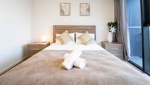 A hotel room with a large bed, two bedside tables, a nightstand, a window with curtains, and two wall-mounted artworks. - Prime Spot: Modern 2BD/2BA with Pool & Parking M1 (South Brisbane)