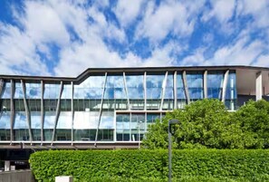 A modern building with a glass facade, a green hedge in the foreground, and a clear blue sky. - Prime Spot: Modern 2BD/2BA with Pool & Parking M1 (South Brisbane)