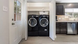 A laundry room with two front-loading washing machines, a white door, and a kitchen area with dark cabinets and a stainless steel dishwasher. - Chic Contemporary Home w/ Park + Garage/Pets ok (Conroe)