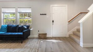 A living room with a dark-colored sofa, a white door, and a staircase. - Chic Contemporary Home w/ Park + Garage/Pets ok (Conroe)