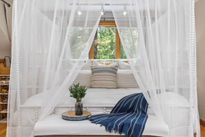 Una cama con dosel, vista de árboles a través de una ventana y una pequeña mesa redonda con una planta.