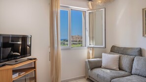 Ein Zimmer mit Fernseher auf einem Standfuß, einer Couch, einem Fenster mit Blick auf eine Hügel- und Meereslandschaft sowie einem Vorhang.