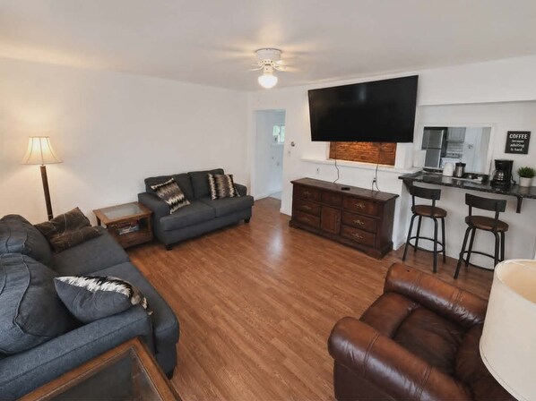 A living room with a sofa, a coffee table, a TV, a kitchen area, and a dining table with chairs. - Pittsburgh Fan Favorite (Homestead)