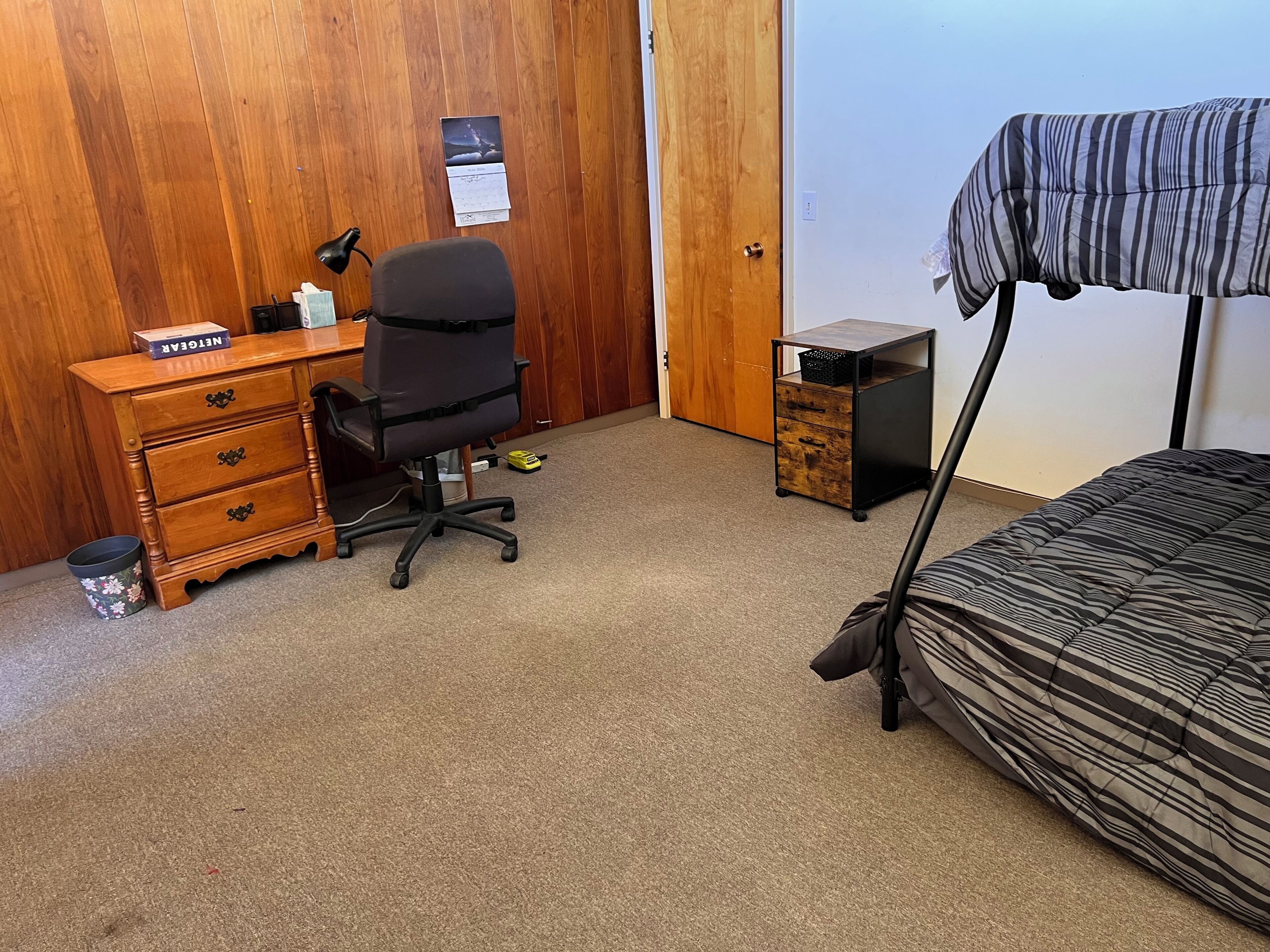 A bedroom with a wooden wardrobe, a desk with a chair, a small cabinet, and a bed with a striped blanket.