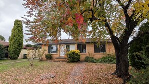 Une maison d’habitation avec une façade en briques, un jardin devant la maison avec un chemin, et un grand arbre aux feuilles d’automne.