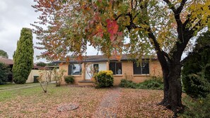 A residential house with a brick exterior, a front yard with a pathway, and a large tree with autumn leaves.