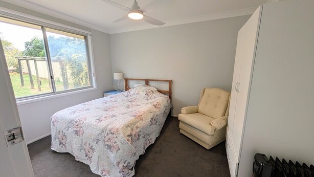 Une chambre à coucher comprenant un lit, une chaise, une table de chevet, un ventilateur de plafond et une fenêtre.