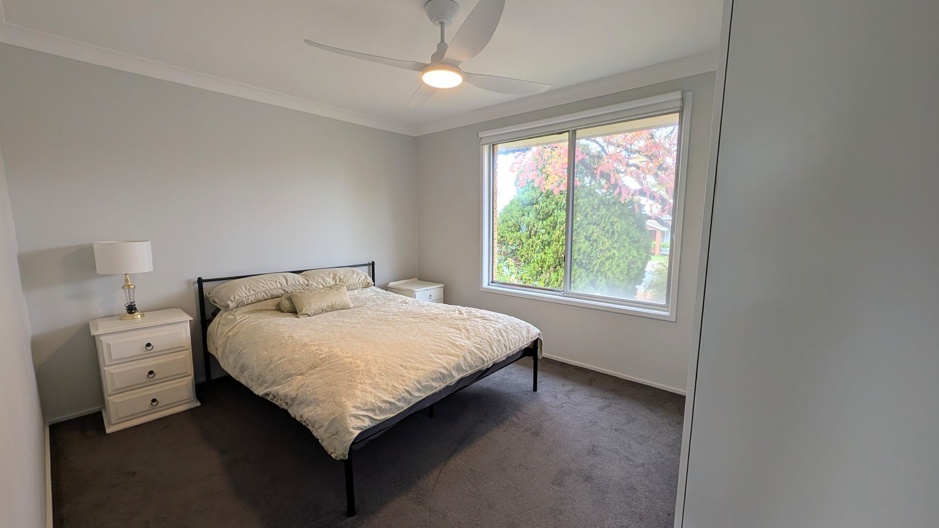 A bedroom with a bed, a nightstand, a lamp, a ceiling fan, and a window with a view of trees.