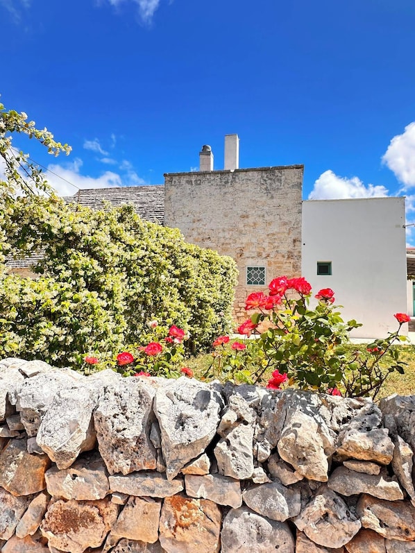 Un muro de piedra, un edificio con chimenea y flores rojas en primer plano.