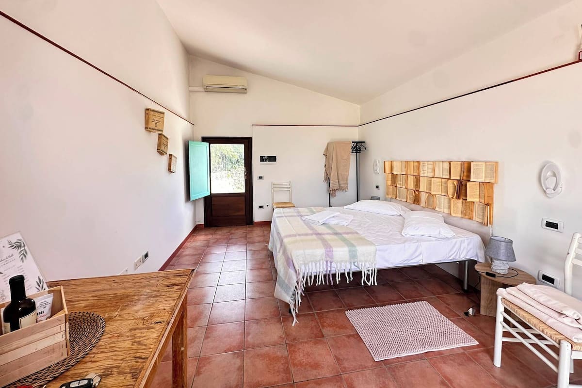 Un dormitorio con una cama, una mesa de madera con una botella, un banco, un tapete y un cabecero con libros.