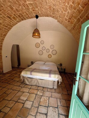 A bedroom with a stone floor, a bed with a plaid blanket, a hanging lamp, and a wall with circular decorations. - Giardino dei Trulli Cummerse (Castellana Grotte)
