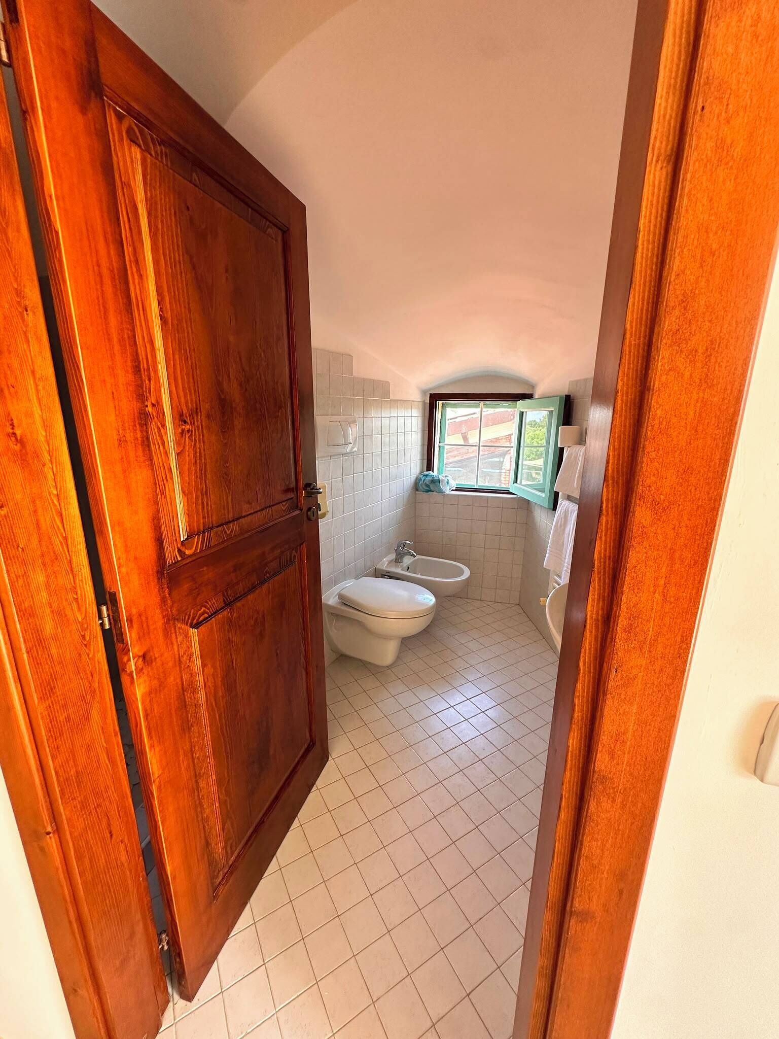 Un baño con inodoro, bidé y lavamanos, suelo de baldosas y una ventana que deja entrar luz natural.