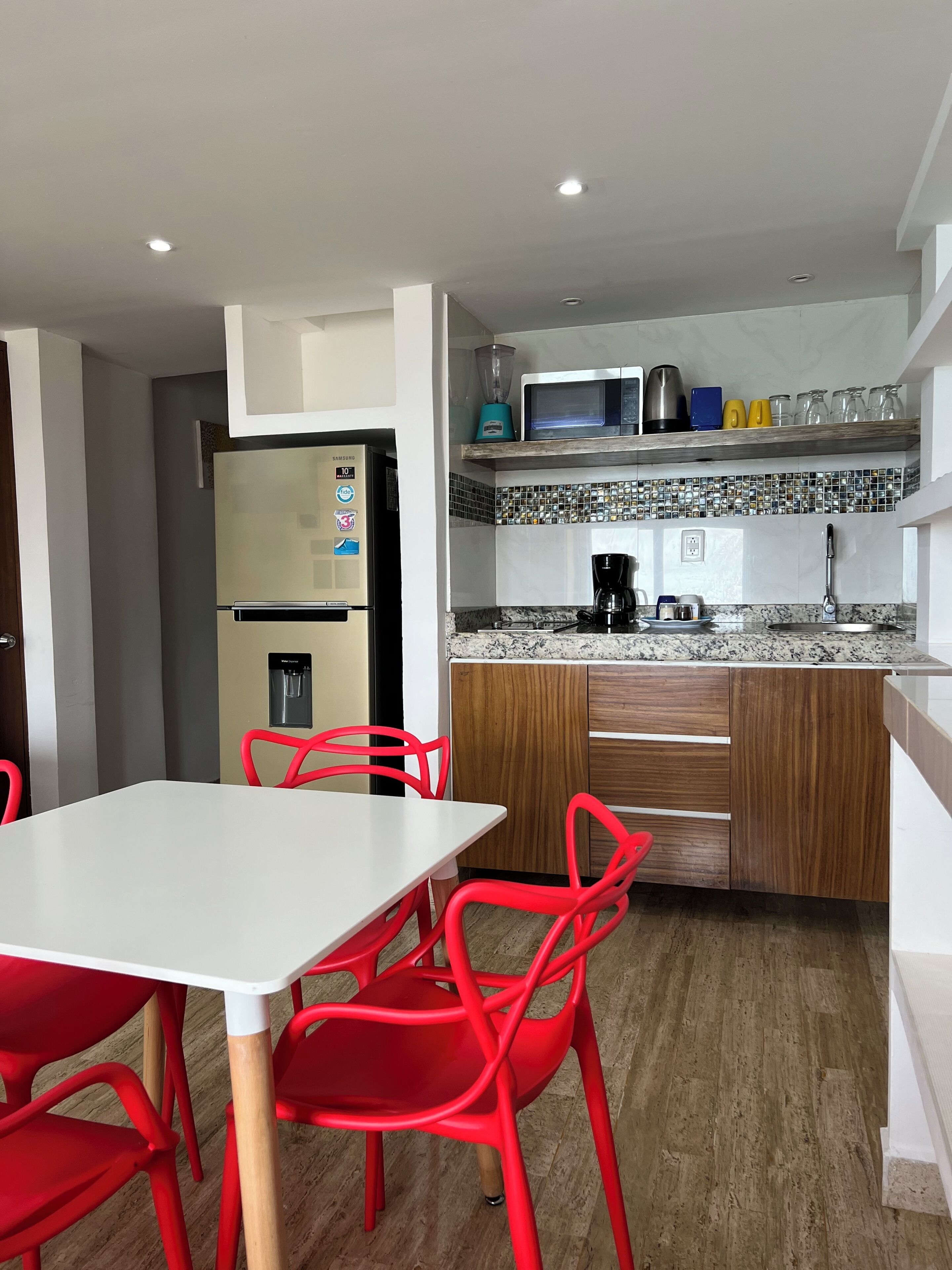 Una cocina moderna con refrigerador, microondas y cafetera. Cuenta con una mesa de comedor con sillas rojas y un dispensador de agua.
