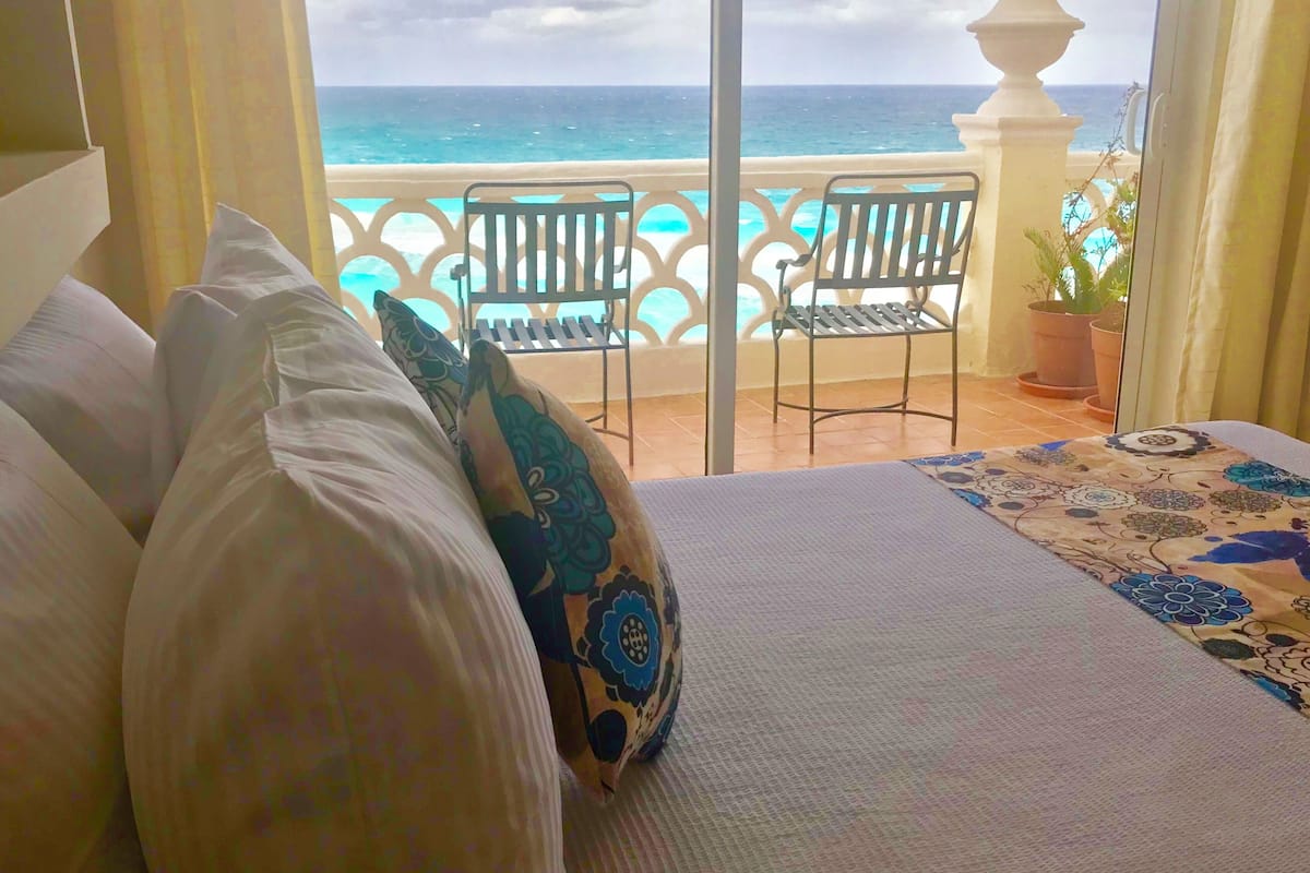 A bed with pillows, a view of the ocean, and two chairs on a balcony.