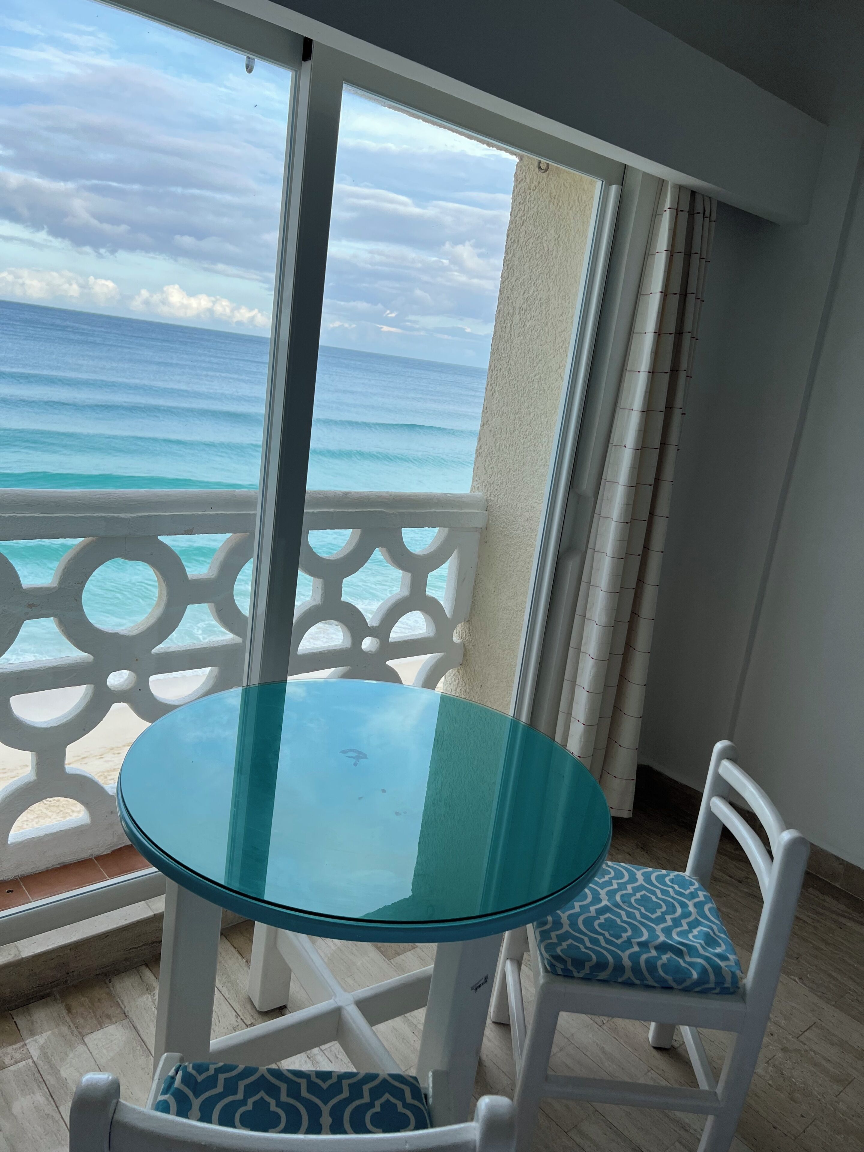 Un balcon avec une table et des chaises en verre, donnant sur une plage où l’on peut admirer les vagues.