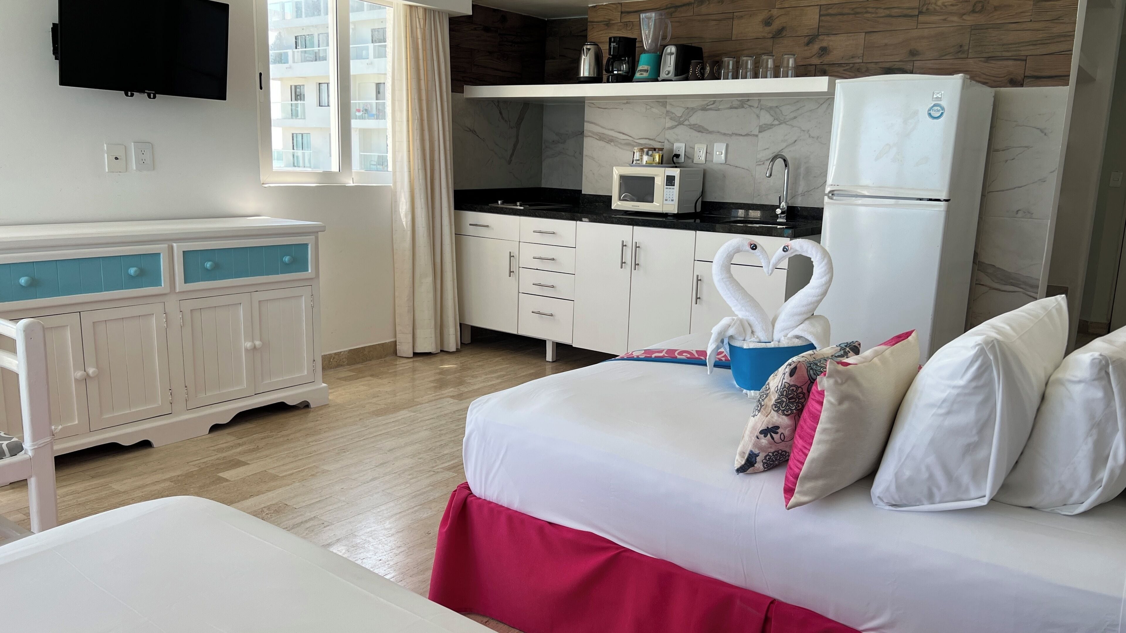 A hotel room with a bed, a kitchenette, a TV, and a window with curtains.