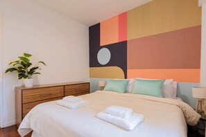 A hotel room with a bed, a dresser, a potted plant, and a colorful abstract wall mural. - MOT Rentals + Junin - 2 Bd (Buenos Aires)