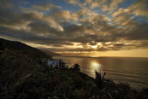 Un paisaje costero con una casa que domina el mar, cielo despejado y un atardecer.