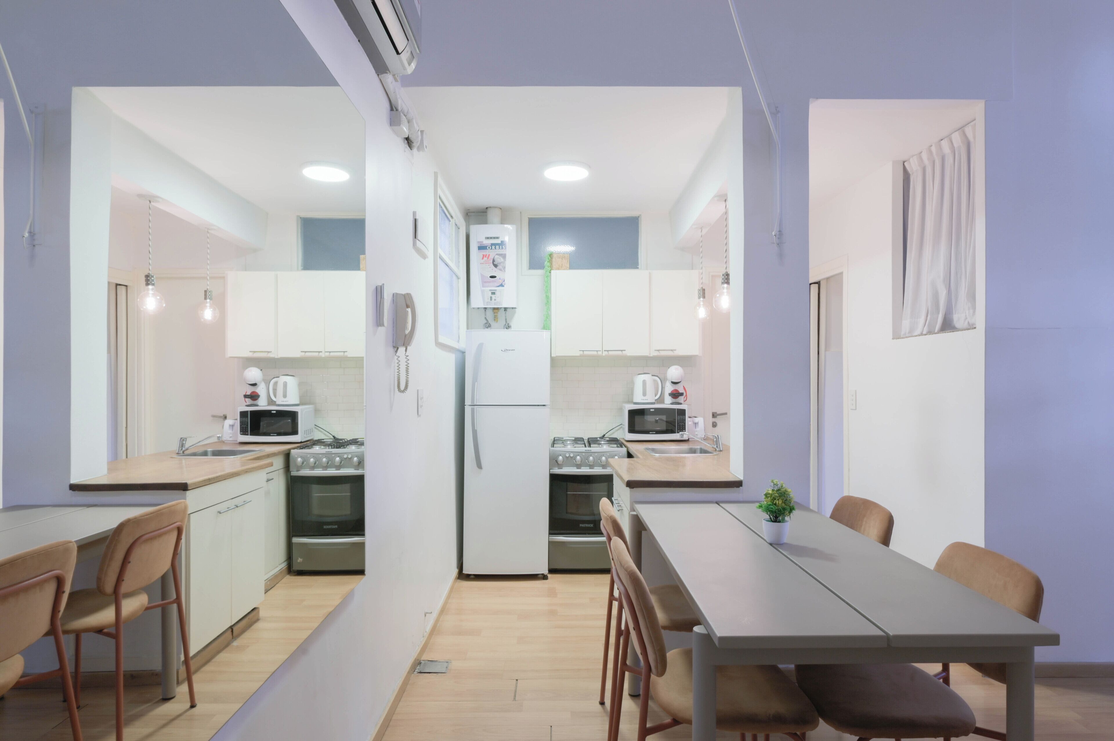 Una cocina compacta con gabinetes blancos, un refrigerador, una estufa y un microondas. Hay una mesa de comedor con sillas y una pequeña planta en maceta.