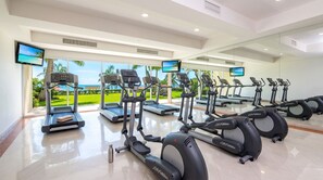 Ein Fitnessraum mit Laufbändern und Crosstrainern, großen Fenstern und Blick auf einen Strand.