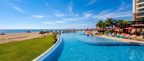 Piscina com vista para a praia e oceano, rodeada por espreguiçadeiras e guarda-sóis.