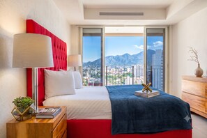 A hotel room with a bed, a nightstand, a lamp, a plant, and a view of mountains. - Gran Venetian Resort Stay Oceanfront by Monyxbnb (Puerto Vallarta)