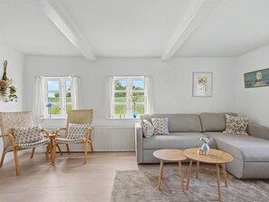 Ein Wohnzimmer mit grauem Sofa, zwei Holzstühlen, einem kleinen runden Tisch und einem Fenster mit Blick auf Grünflächen.