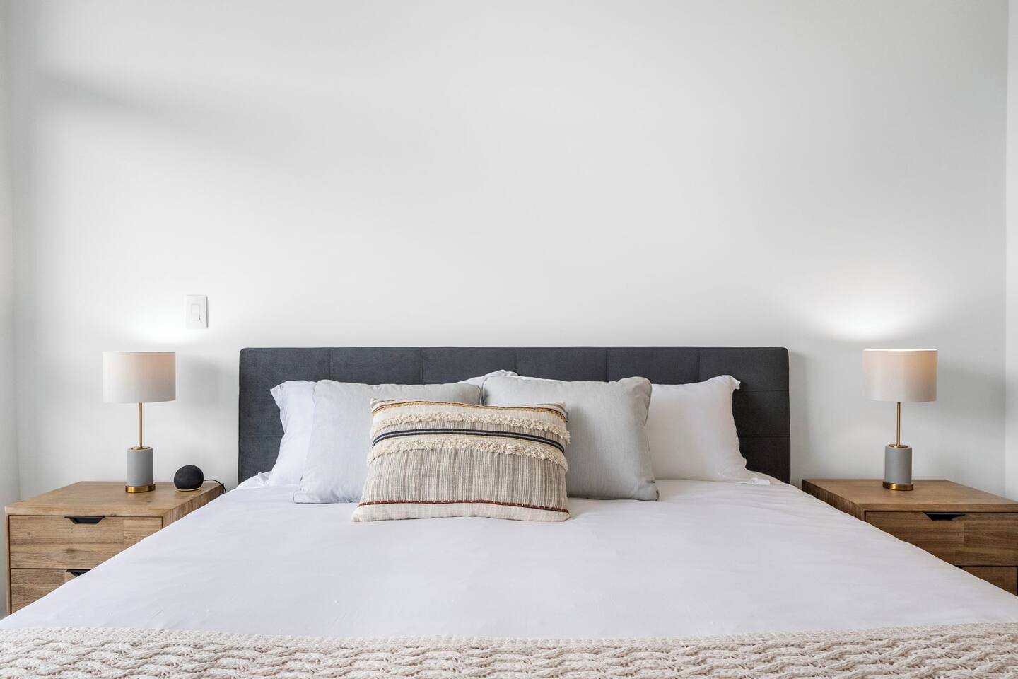 Un lit bien fait, avec une tête de lit grise, deux tables de chevet dotées de lampes et un plaid à motifs.