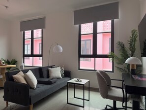 Ein modernes Wohnzimmer mit einem grauen Sofa, einem kleinen Tisch, einem Sessel und großen Fenstern mit Blick auf rosafarbene Gebäude.
