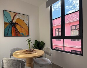 Ein Zimmer mit einem runden Tisch, Stühlen, einem Gemälde, einer Topfpflanze und einem Fenster mit Blick auf ein rosafarbenes Gebäude.
