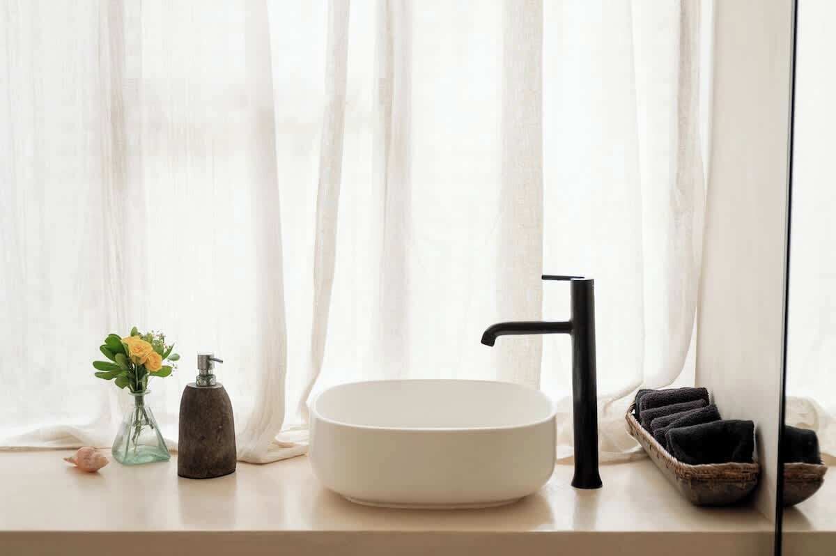 Un bagno con lavandino bianco, rubinetto nero, un piccolo vaso con fiori gialli, un dispenser per sapone e un cesto con asciugamani.