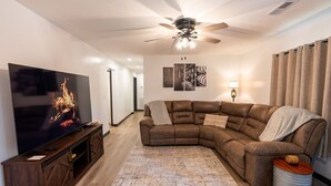 Un salon comprenant un canapé d’angle, un téléviseur à écran plat, un ventilateur de plafond et un tapis à motifs.