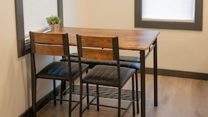 Un coin repas avec une table en bois et quatre chaises, deux fenêtres équipées de stores et une petite plante posée sur la table.