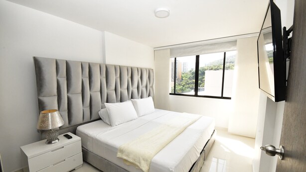 A modern bedroom with a large bed, a nightstand, a lamp, and a TV mounted on the wall. - Stylish 12th Floor Apartment in the Heart of El Peñón (Cali)