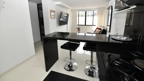 A modern kitchen with a black countertop, two bar stools, a stainless steel range hood, and a flat-screen TV mounted on the wall. - Stylish 12th Floor Apartment in the Heart of El Peñón (Cali)