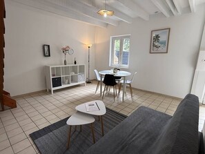 Ett vardagsrum med en soffa, ett matbord med stolar, ett vitt skåp, en golvlampa och ett fönster med utsikt.