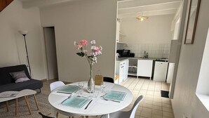 A modern living room with a round dining table set for four, a sofa, a vase with flowers, and a kitchen in the background. - NEW LISTING, Domaine de KER-PAULT, between St Cast and Matignon, 3-room cottage 5 minutes from... (MATIGNON)