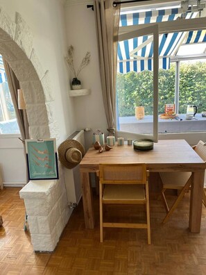 Uma mesa de madeira com cadeiras, um quadro emoldurado, uma janela com vista para o verde e um toldo listrado.