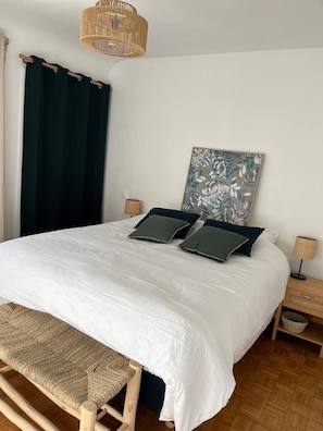 Ein Bett mit weißer Decke, zwei dunklen Kissen, eine gewebte Bank, ein gewebter Nachttisch, eine gewebte Lampe und ein an der Wand befestigter Bilderrahmen.