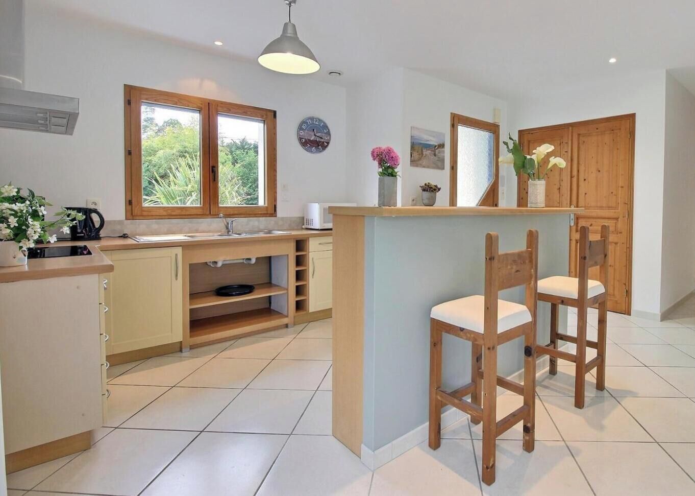 A modern kitchen with wooden cabinets, a built-in island with bar stools, a window with a view, and a wall clock.