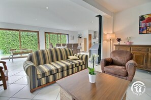 Ein modernes Wohnzimmer mit einer Couch, Sesseln und einem Couchtisch. Es gibt einen Kamin, einen Essbereich mit Tisch und Stühlen sowie große Fenster mit Blick auf Grünflächen.