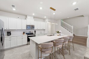A modern kitchen with white cabinets, stainless steel appliances, a central island with bar stools, and a staircase leading to another level. - 5BR Home with Pool & Game Room at Windsor Cay (Clermont)
