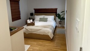 Een smalle gang die leidt naar een slaapkamer met een bed, nachtkastje en een kamerplant.