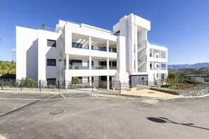 Ein modernes, mehrgeschossiges Gebäude mit Balkonen, einem Parkplatz und einer Bergkulisse.