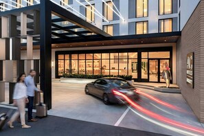 Entrada moderna de hotel con fachada de vidrio, un coche en movimiento y un carrito de equipaje.
