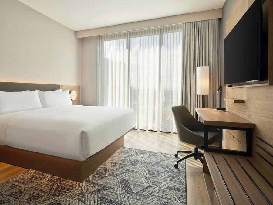 Ein Hotelzimmer mit Bett, Schreibtisch, Stuhl, Fernseher und einem großen Fenster mit Vorhängen.
