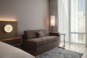 A modern hotel room with a sofa, a bedside table, a lamp, and a view of the outdoors through sheer curtains.