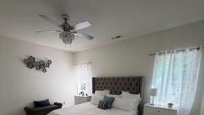 Quarto com cama grande, mesa de cabeceira, cadeira, ventilador de teto e um objeto decorativo na parede.