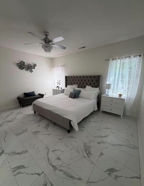 A bedroom with a large bed, a nightstand, a chair, a ceiling fan, and a decorative wall piece.
