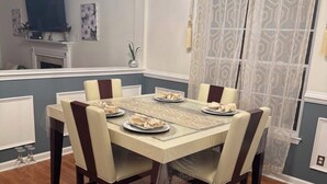Uma sala de jantar com uma mesa posta para seis pessoas, rodeada por cadeiras, um lustre, uma televisão e uma cortina estampada.